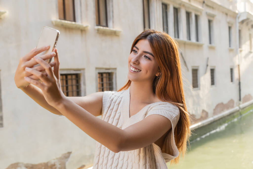 Venedik 'teki bulanık binanın yakınındaki akıllı telefondan selfie çeken neşeli kadın. - Fotoğraf, Görsel
