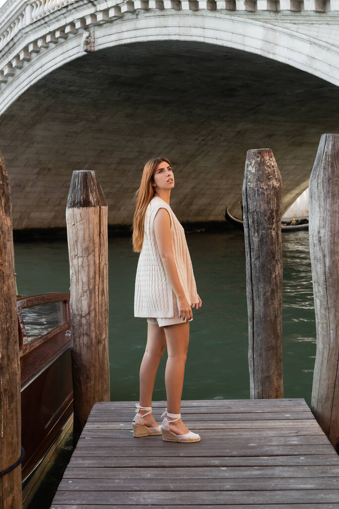 donna magra in maglieria estiva e sandali con zeppa che distoglie lo sguardo sul molo di legno a Venezia - Foto, immagini