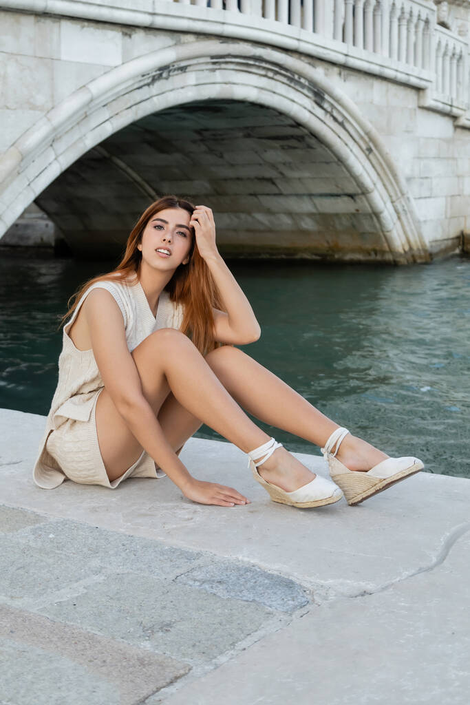 Venedik Kanalı 'nın yanında otururken takoz sandaletli güzel bir kadın kızıl saçlarını düzeltiyor. - Fotoğraf, Görsel