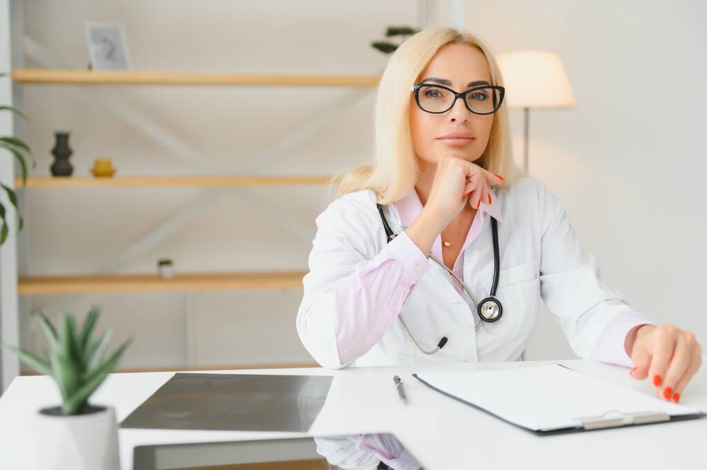 Портрет женщины-врача средних лет, сидящей за столом и работающей в офисе врача. - Фото, изображение