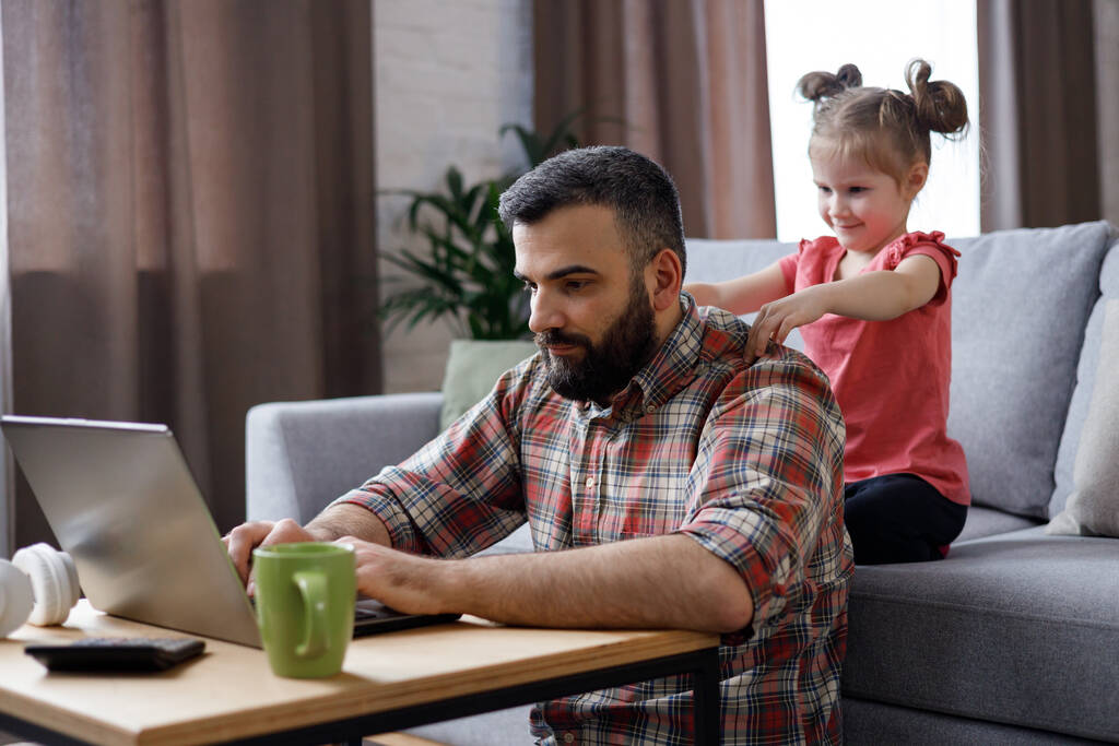 Счастливый семейный портрет. Молодой красивый мужчина работает дома за ноутбуком, фокусирует внимание на экране ноутбука, пока его маленькая дочь пытается массировать его плечи. Семейный уход и поддержка. - Фото, изображение