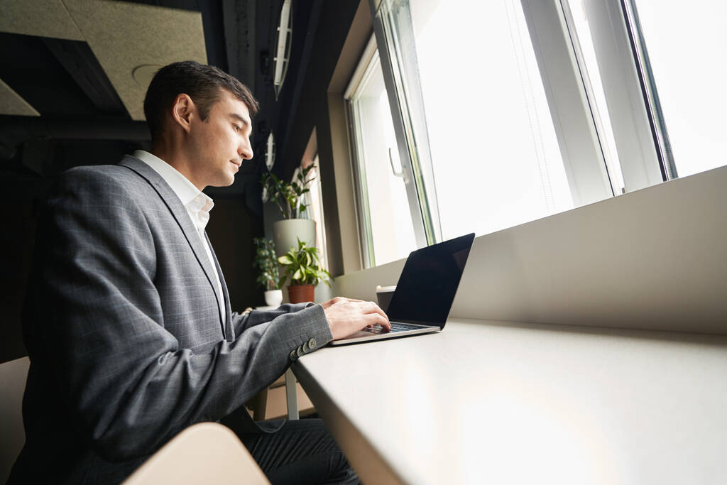 Vista lateral del empleado de oficina que trabaja en el teclado del ordenador portátil mientras está sentado cerca de la ventana - Foto, imagen