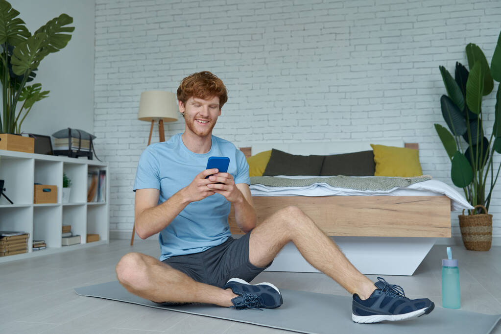 Jóképű férfi sportruhában okos telefont használ, miközben otthon ül a gyúrószőnyegen. - Fotó, kép