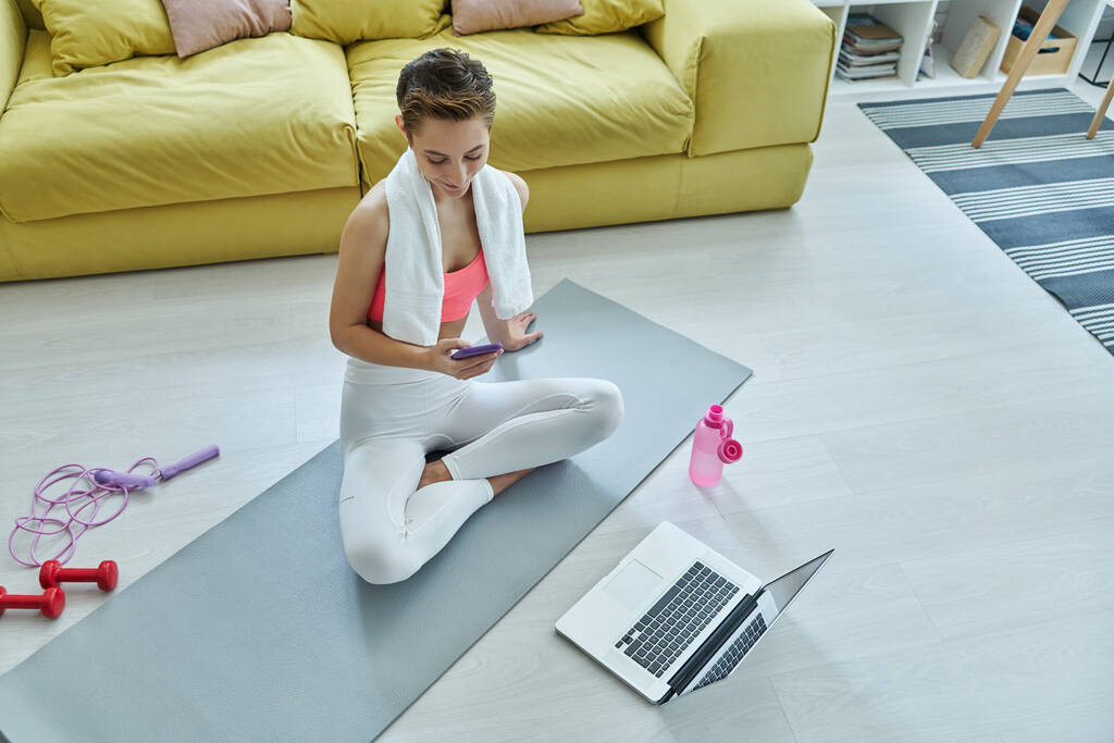 Красива жінка в спортивному одязі, використовуючи мобільний телефон, сидячи на тренувальному килимку вдома
 - Фото, зображення