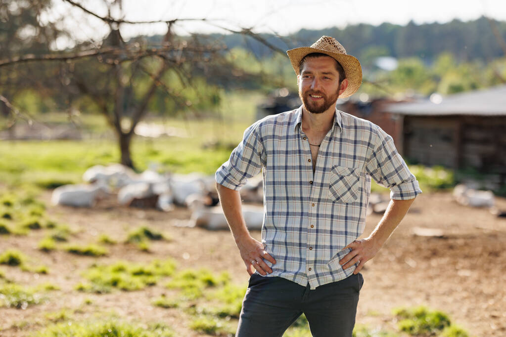 Egy fiatal, fehér, jóképű, szakállas férfi farmer portréja, aki a kamerába mosolyog. Boldog farmer szalmakalapban az ökotelepen. Kecsketejtermékek ipari előállítása. Szarvasmarha-tenyésztés. - Fotó, kép