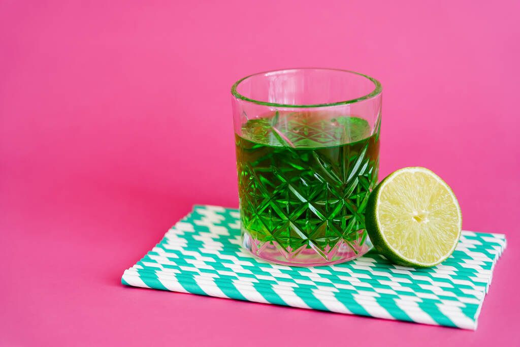 glass of green alcohol drink on striped paper straws near sliced lime on pink - Foto, Imagem