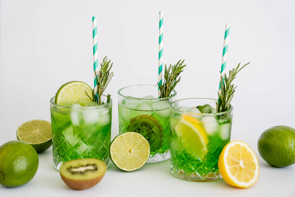 граненые бокалы с холодным зеленым коктейлем, соломинками и фруктами на белом  - Фото, изображение