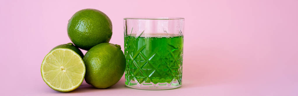 πράσινο αλκοόλ ποτό σε ποτήρι κοντά φρέσκα και οργανικά λάιμ σε ροζ, banner - Φωτογραφία, εικόνα