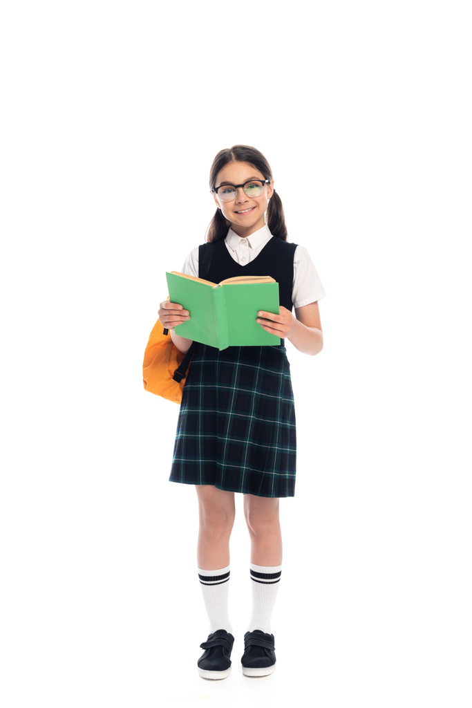 Teljes hossza iskolás szemüveges holding book és mosolygós kamera fehér háttér - Fotó, kép