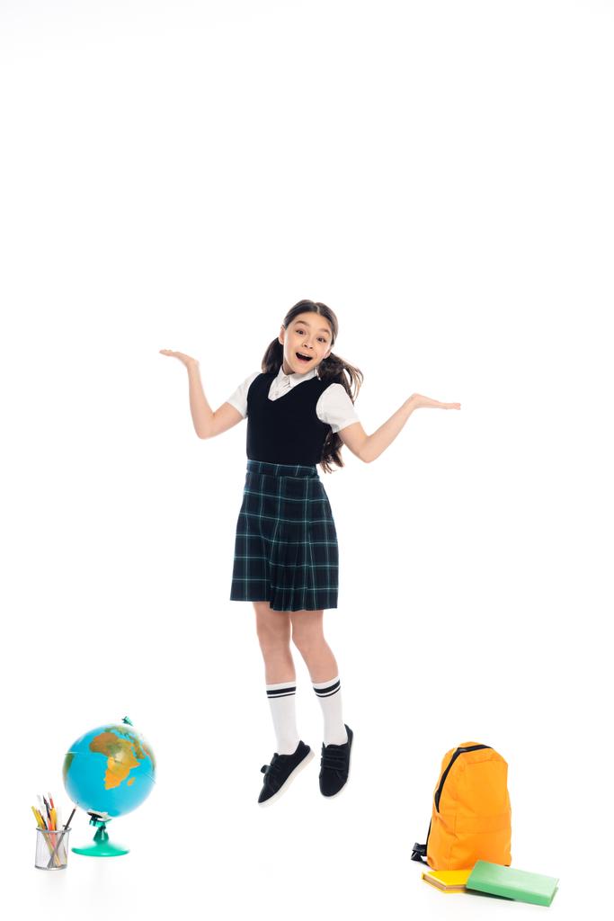 Positive schoolkid pointing with hands while jumping near globe and books on white background - Photo, Image