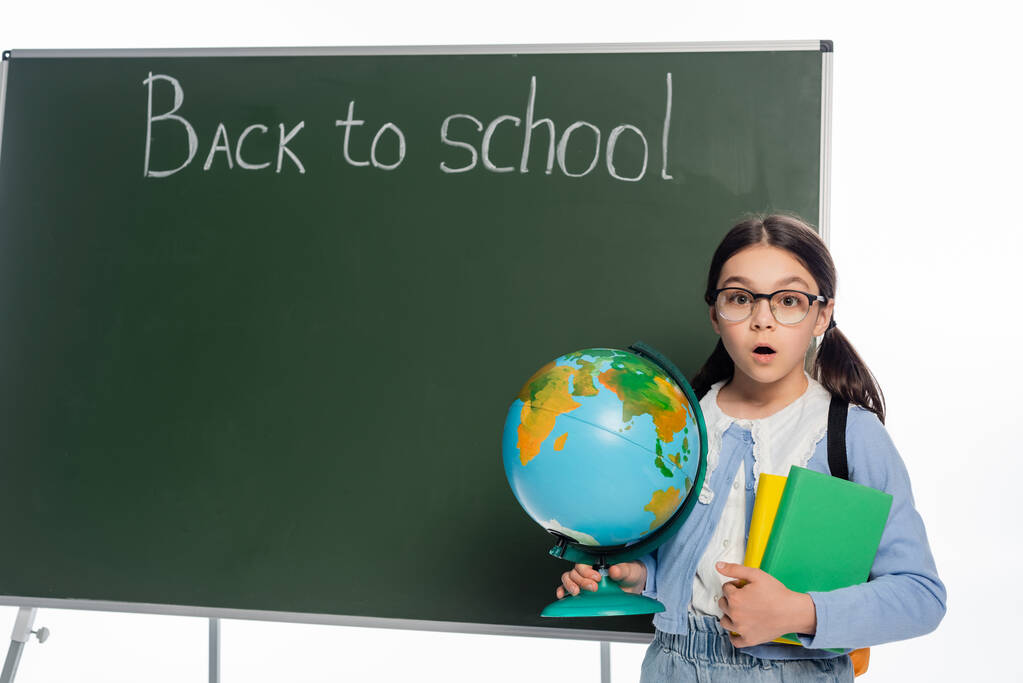Choqué écolier tenant des livres et globe près de tableau noir avec retour à l'école lettrage isolé sur blanc  - Photo, image