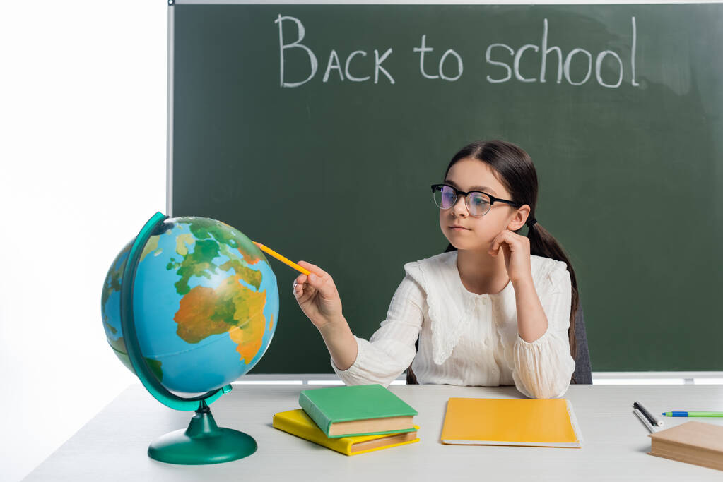 Schulmädchen zeigt auf Globus in der Nähe von Büchern und Kreidetafel mit Schulbuchstaben auf weißem Hintergrund  - Foto, Bild