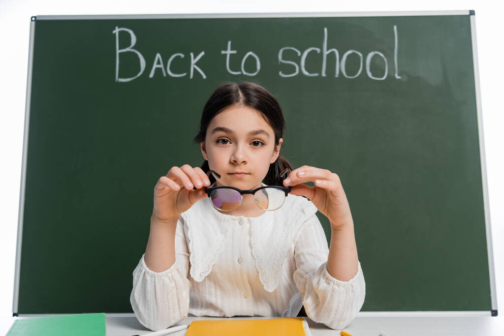 Школярка тримає окуляри поблизу блокнота та крейди зі спиною до шкільного написання ізольовані на білому
  - Фото, зображення
