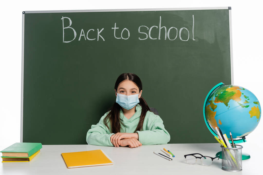 Školák v lékařské masce sedí v blízkosti knih a tabuli s zpět do školy písmo izolované na bílém  - Fotografie, Obrázek