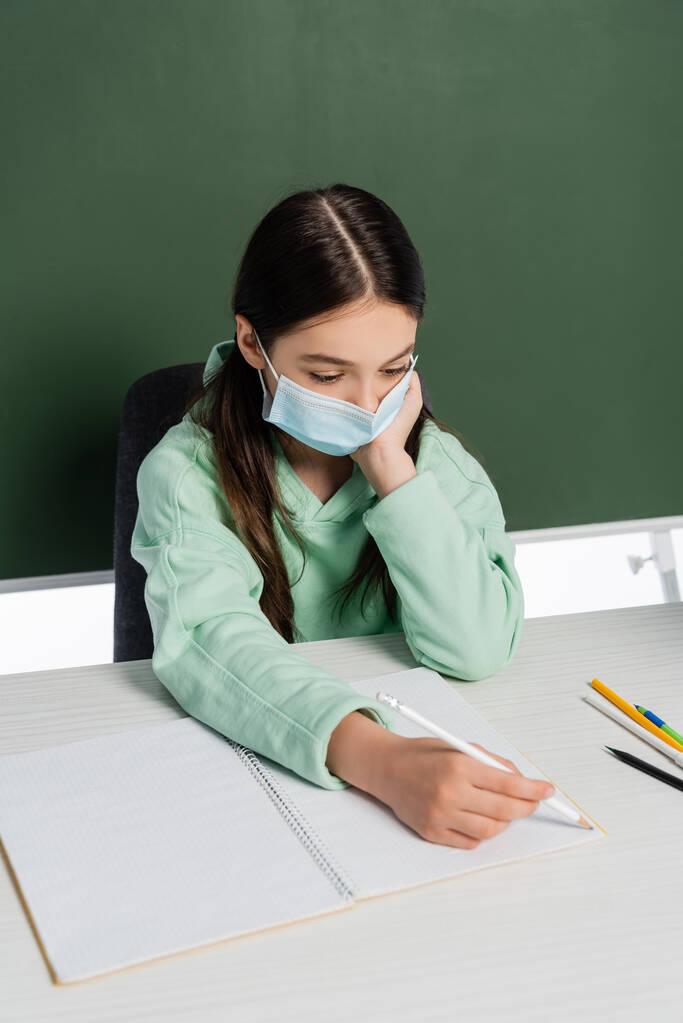 Écolière en masque médical écrivant sur carnet près de tableau isolé sur blanc  - Photo, image