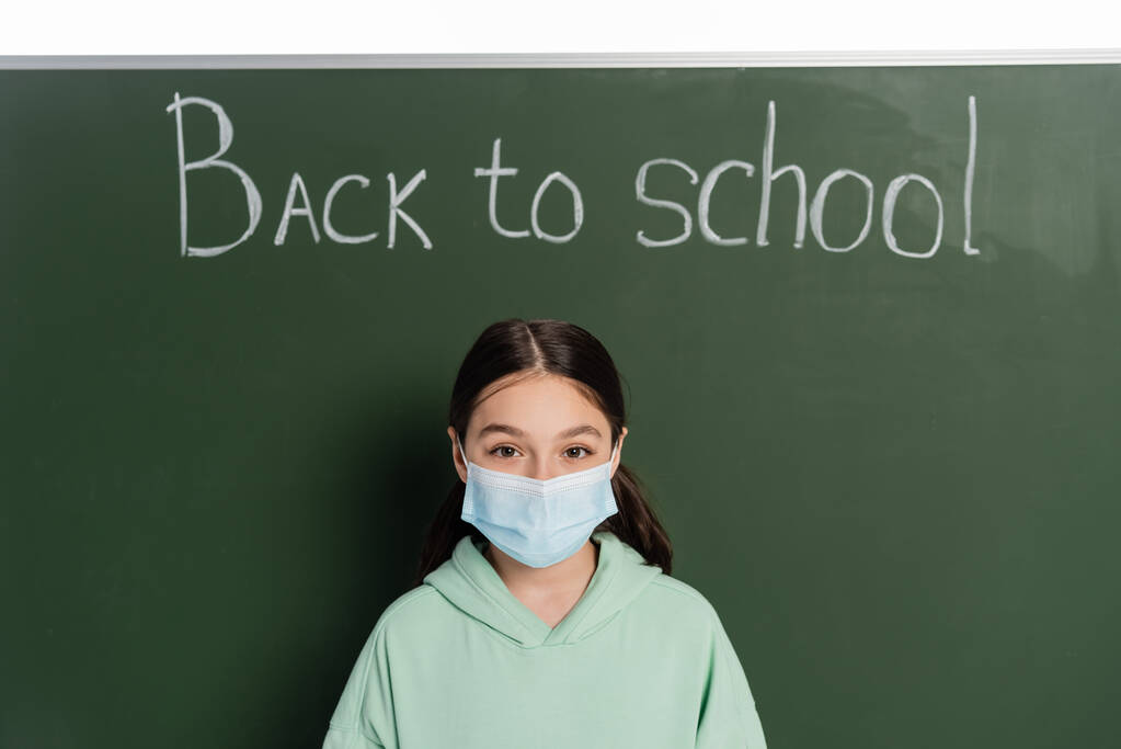 Preteen Schulmädchen in medizinischer Maske blickt in die Kamera in der Nähe der Tafel mit dem Rücken zur Schule Schriftzug isoliert auf weiß  - Foto, Bild
