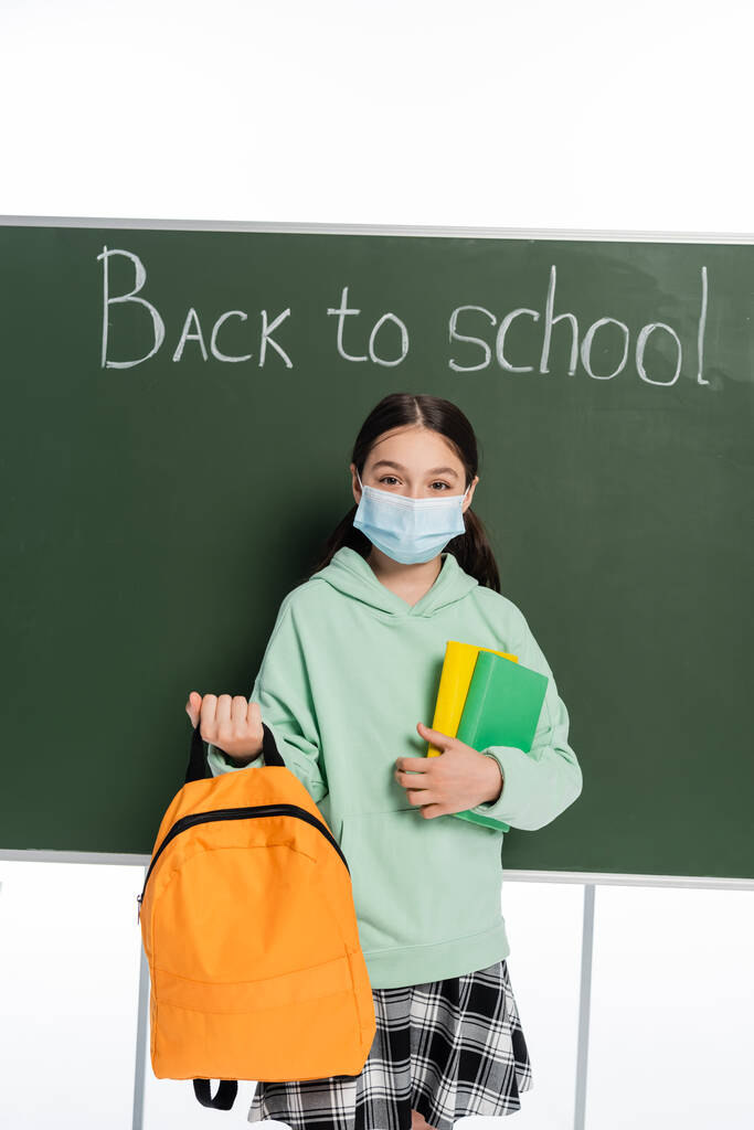 Schulkind in medizinischer Maske mit Büchern und Rucksack in der Nähe von Kreidetafeln mit Schulbuchstaben auf weißem Hintergrund  - Foto, Bild