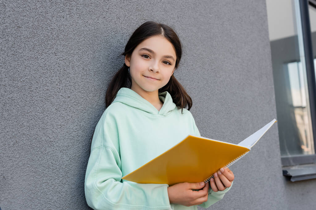 Glimlachend schoolkind met notebook en kijkend naar camera in de buurt van gebouw buiten  - Foto, afbeelding