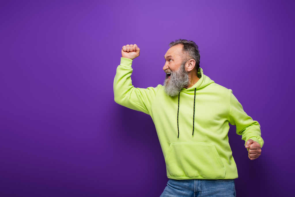 hombre mayor feliz y barbudo con el pelo gris regocijándose mientras mira hacia otro lado en púrpura - Foto, imagen