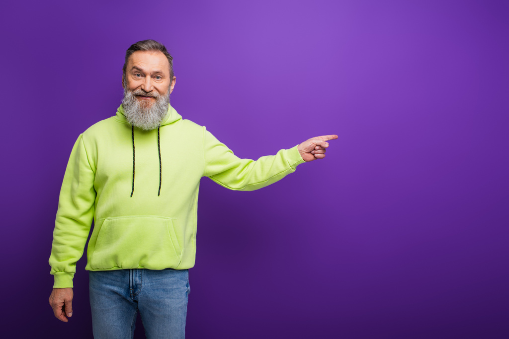 szczęśliwy starszy mężczyzna z brodą i siwymi włosami stojący w zielonej bluzie i wskazujący palcem na fioletowym - Zdjęcie, obraz