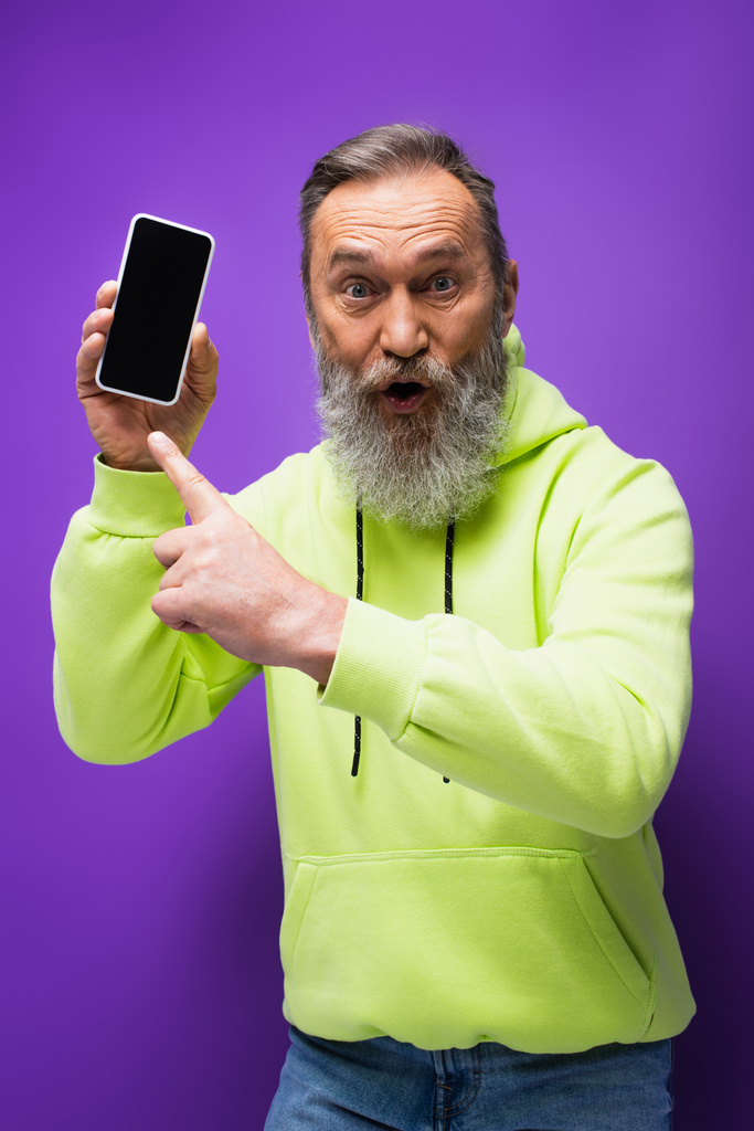 sorprendido hombre mayor con barba y pelo gris apuntando a teléfono inteligente en púrpura  - Foto, imagen