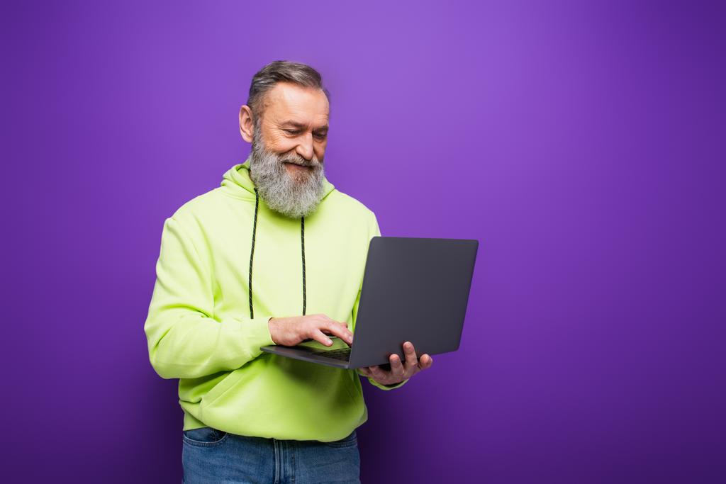 ευτυχισμένος ηλικιωμένος άνδρας με γενειάδα και γκρίζα μαλλιά χρησιμοποιώντας φορητό υπολογιστή σε μωβ  - Φωτογραφία, εικόνα
