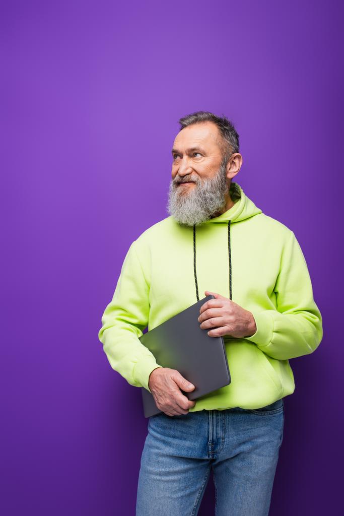 glücklicher älterer Mann mit Bart und grauen Haaren hält Laptop im Stehen und schaut weg auf lila  - Foto, Bild