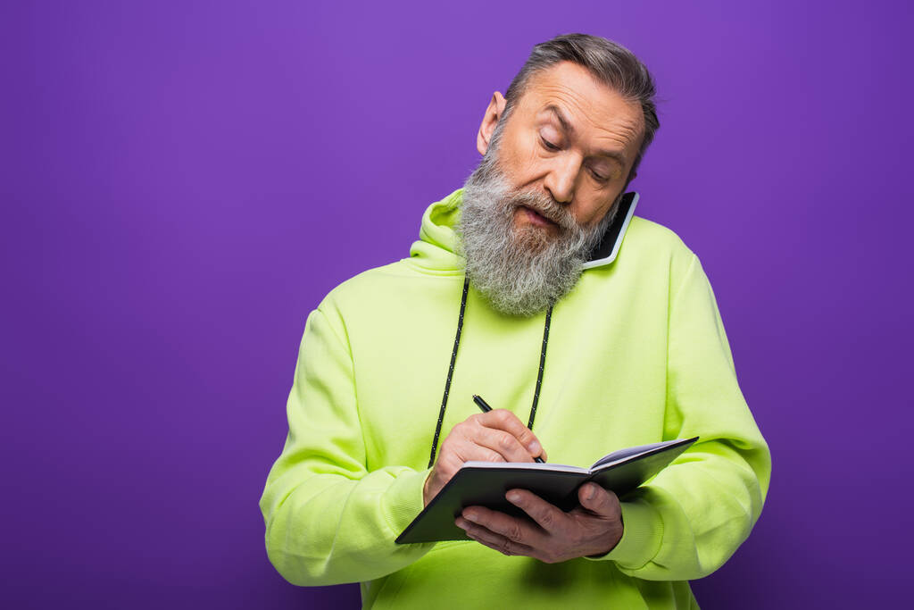 senior man in green hoodie writing on notebook and talking on smartphone on purple  - 写真・画像