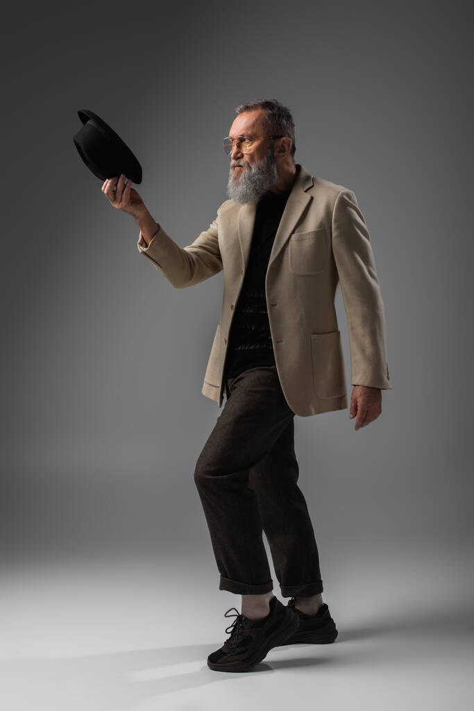 повна довжина бородатого старшого чоловіка в бежевій куртці і окулярах позує з дербі капелюхом на сірому
 - Фото, зображення