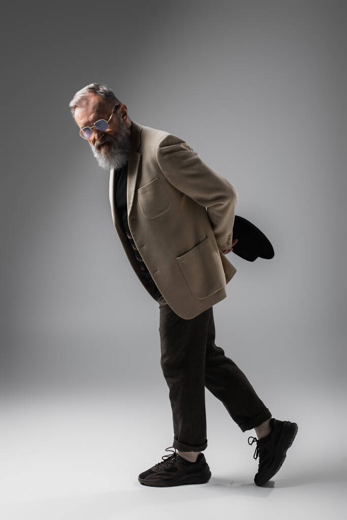 повна довжина стильного старшого чоловіка в бежевому блістері і окулярах позує з дербі капелюхом на сірому
 - Фото, зображення