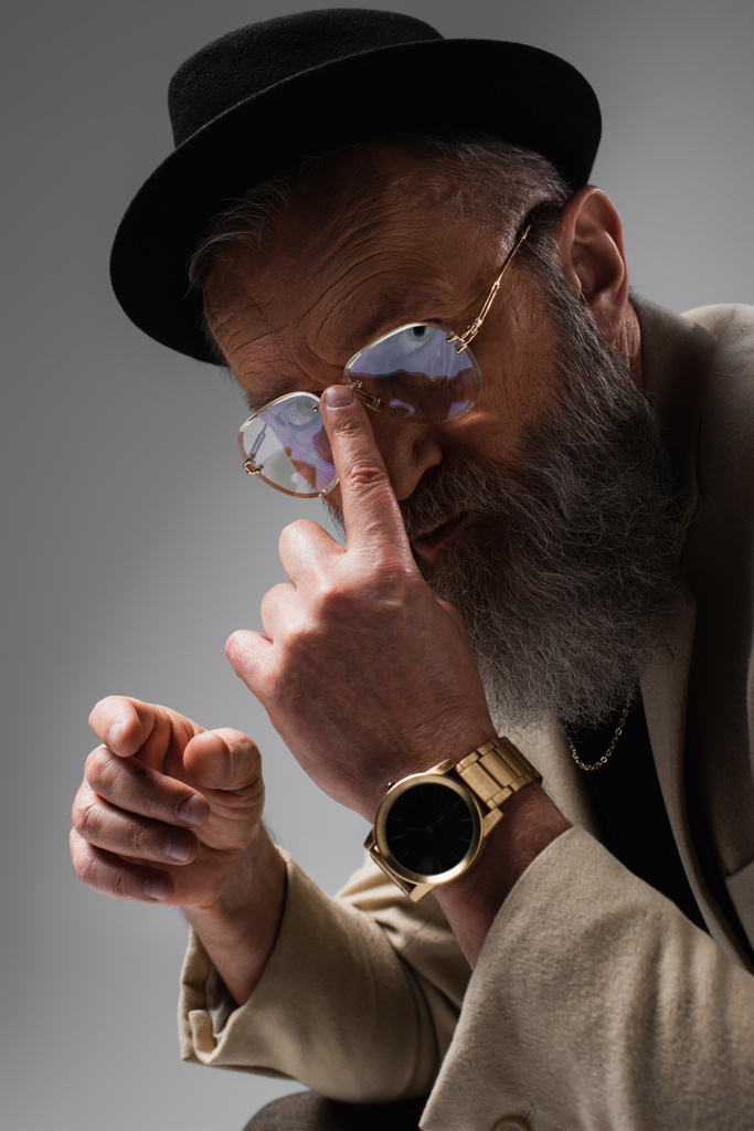 portrait of bearded senior man in stylish beige jacket and derby hat adjusting eyeglasses with finger on grey - Photo, Image
