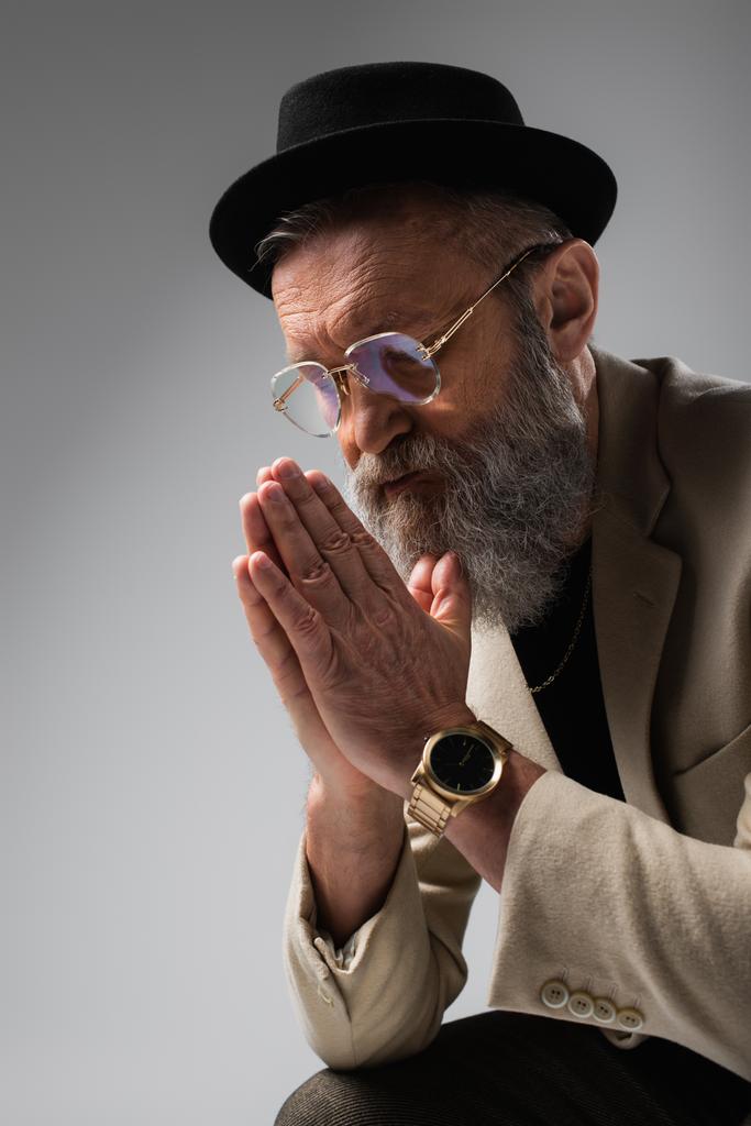 πορτρέτο του γενειοφόρου ηλικιωμένου άνδρα σε κομψό μπεζ σακάκι και καπέλο ντέρμπι με τα χέρια προσεύχεται σε γκρι - Φωτογραφία, εικόνα