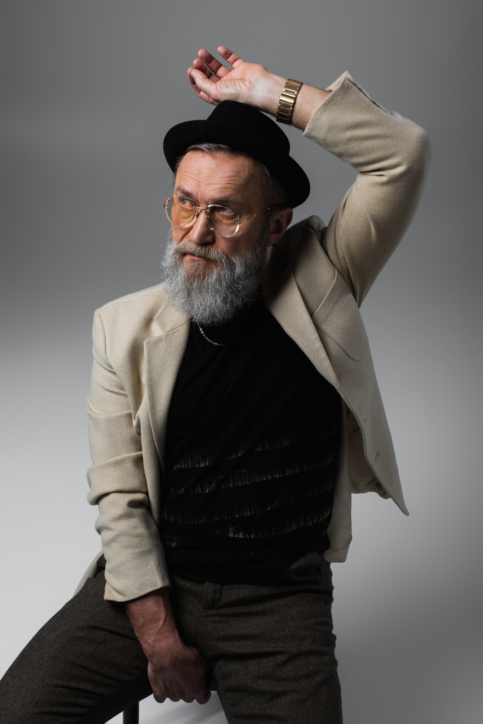 uomo anziano barbuto in cappello derby e occhiali seduti e guardando lontano sul grigio - Foto, immagini