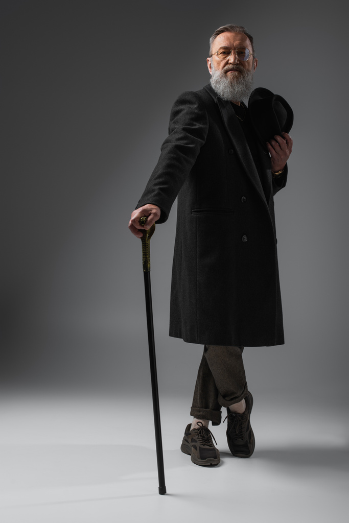 plná délka elegantní senior muž ve stylovém kabátě drží derby klobouk a chodící hůl na šedé - Fotografie, Obrázek
