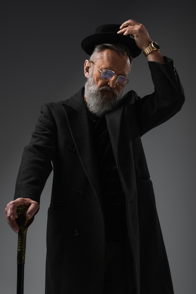 elegant senior man in stylish coat wearing derby hat and holding walking stick on grey - Photo, Image