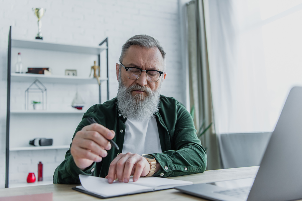 bearded senior man holding pen and looking at notebook near laptop on desk  - Photo, Image