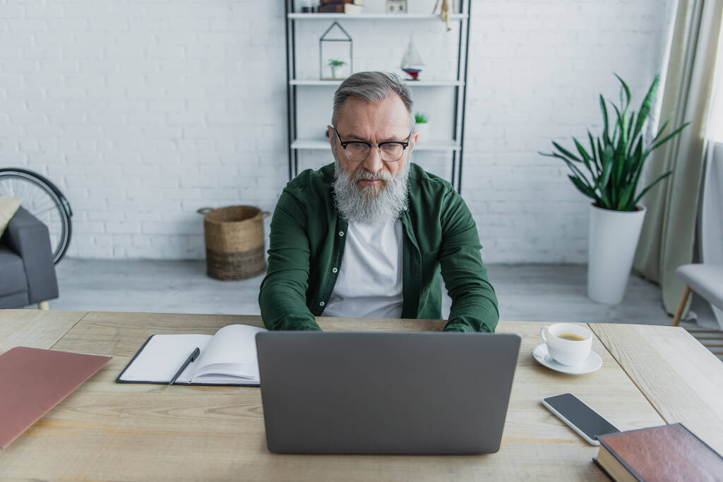 Düşünceli ve sakallı gözlüklü yaşlı adam evde çalışırken laptop kullanıyor.  - Fotoğraf, Görsel