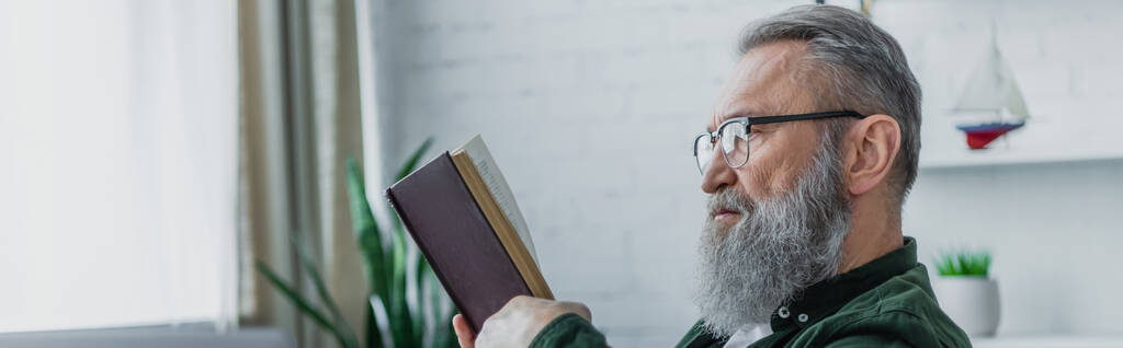 γενειοφόρος ηλικιωμένος άνδρας με γυαλιά ηλίου που διαβάζει βιβλίο στο σπίτι, πανό - Φωτογραφία, εικόνα