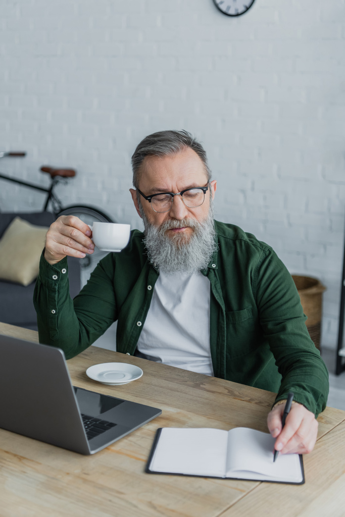 uomo anziano barbuto in occhiali con in mano una tazza di caffè e scrittura vicino al computer portatile mentre si lavora da casa  - Foto, immagini