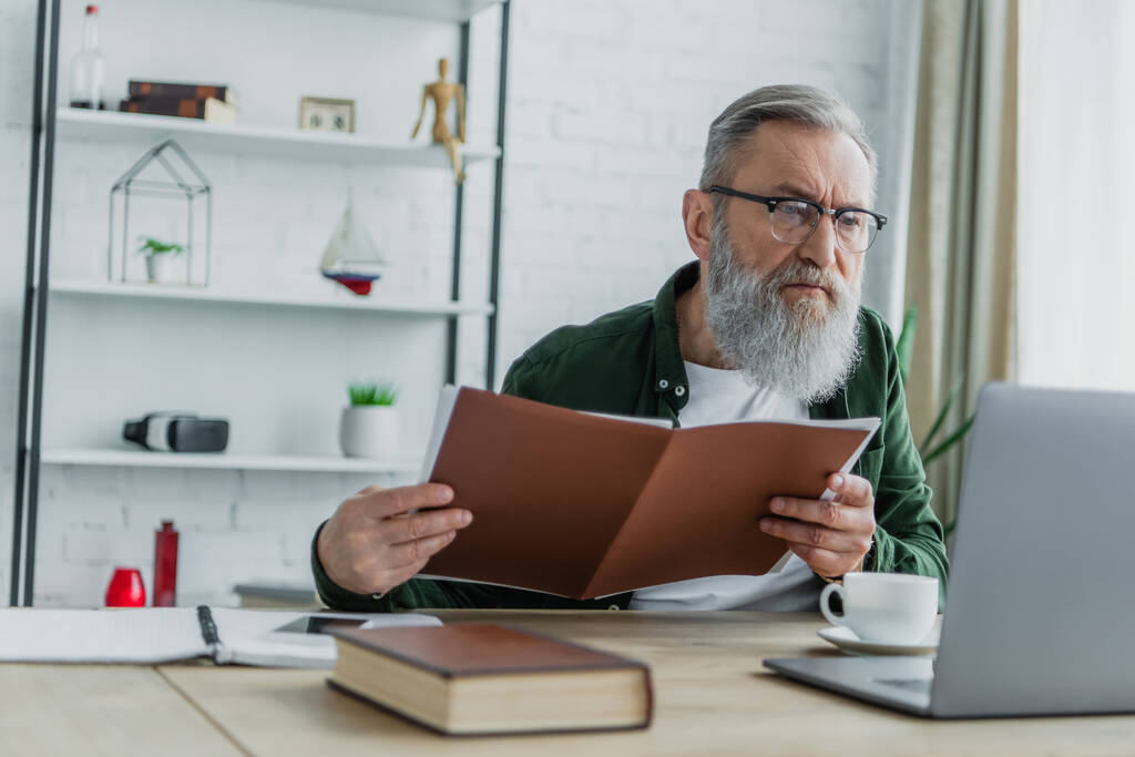 bärtiger älterer Mann mit Brille hält Ordner in der Hand und blickt auf Laptop auf Schreibtisch - Foto, Bild