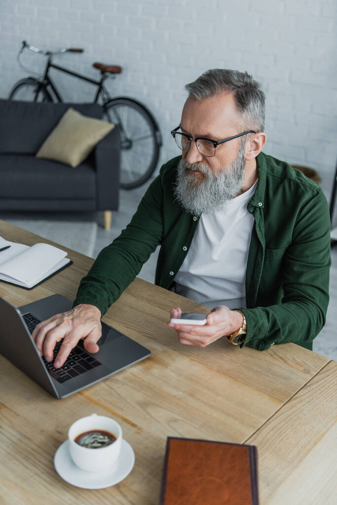 hombre mayor barbudo en gafas con teléfono inteligente mientras usa el ordenador portátil en casa - Foto, imagen
