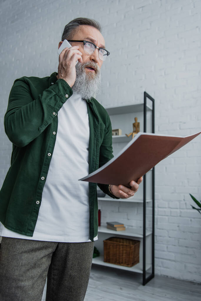 senior businessman with beard in eyeglasses standing with folder while talking on smartphone  - 写真・画像