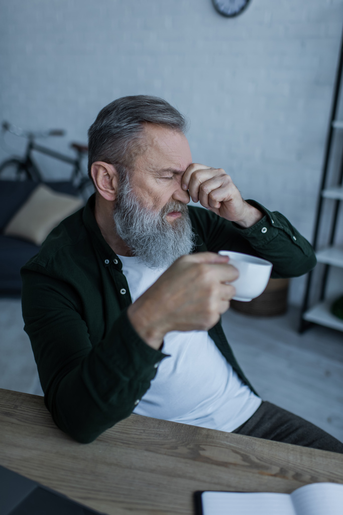 Yorgun sakallı ve yaşlı bir adam, kapalı gözlere dokunurken bir fincan kahve tutuyor. - Fotoğraf, Görsel