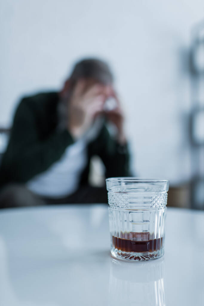egy pohár whisky a fehér dohányzóasztalon a homályos, krízisben szenvedő férfi közelében  - Fotó, kép