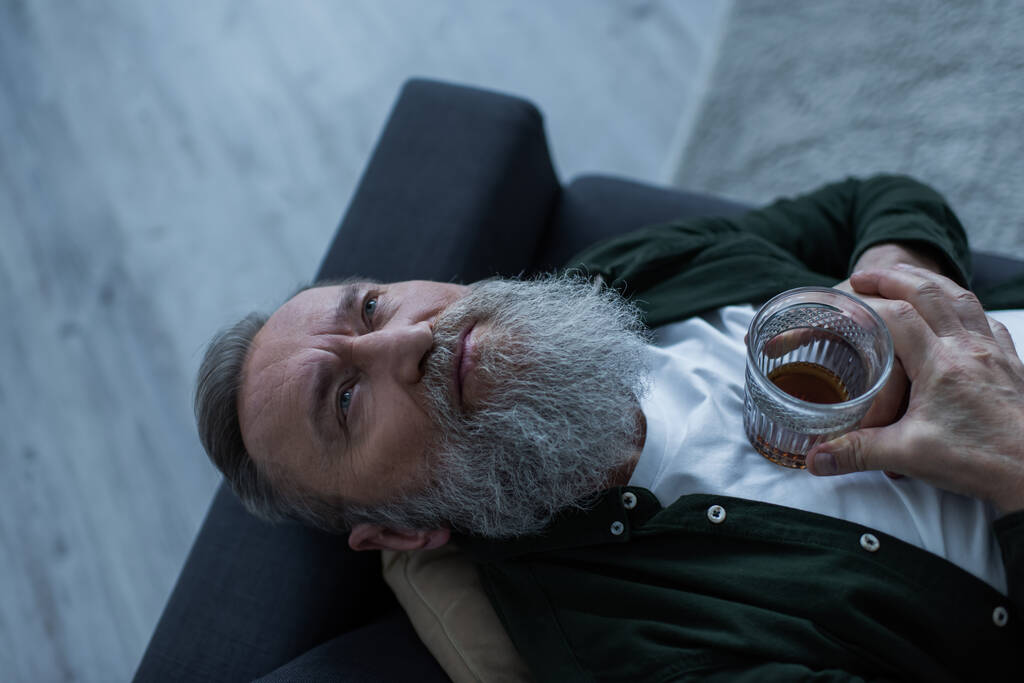 top view of disappointed senior man with beard lying on couch with glass of whiskey while suffering crisis - 写真・画像