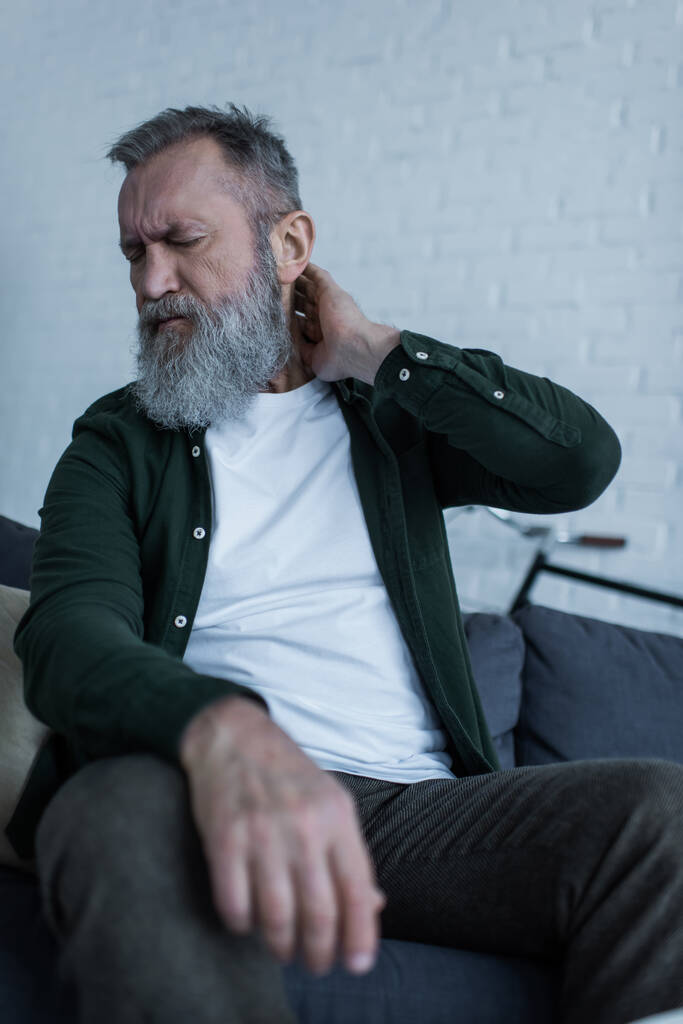 upset senior man with beard and closed eyes sitting on couch and having neck pain - 写真・画像