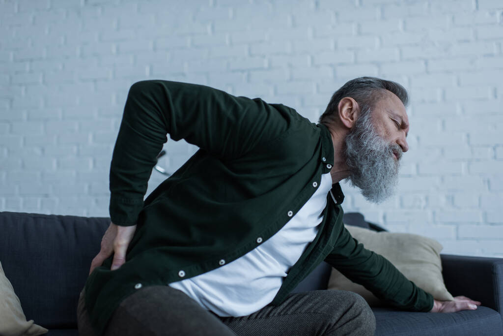 бородатый мужчина с бородой сидит на диване и трогает спину, страдая от боли  - Фото, изображение