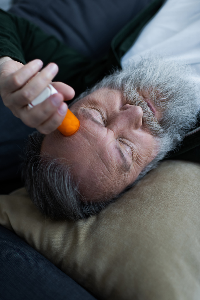 nešťastný senior muž drží láhev s antidepresivními léky, zatímco leží na gauči v obývacím pokoji - Fotografie, Obrázek