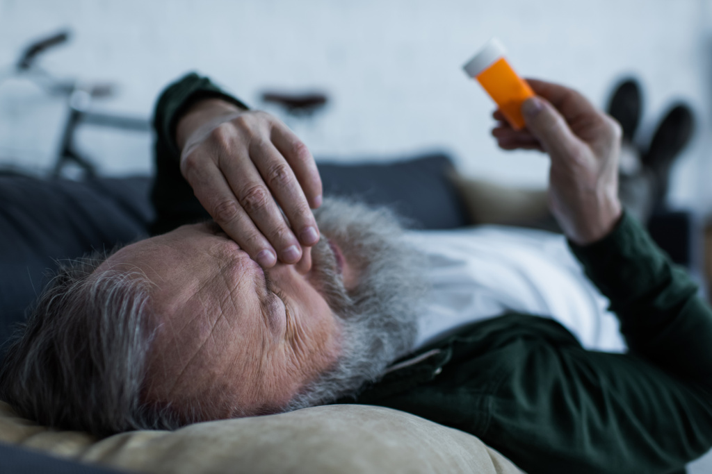 depressed senior man holding bottle with medication while lying on couch in living room - Photo, Image