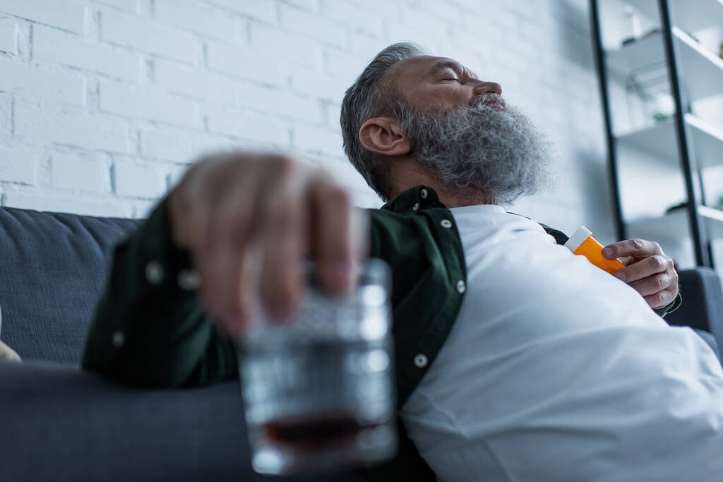 brodaty starszy mężczyzna z siwymi włosami trzymający butelkę z lekami i szklanką whisky  - Zdjęcie, obraz